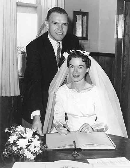 Smiling Happy Bride Sits Signing Wedding Registry Book With Groom Standing Behind Stylish Classy Couple 1959