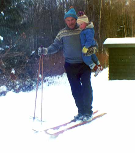 Grandfather Outdoors In Snow Standing On Wood Skis Bamboo Poles Wearing Touque Sweater Holding Grandson 1970