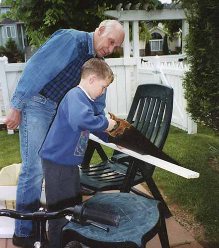 Grandfather Holds Board On Chair Arms Outside While Teaching Young Grandson To Use Wood Hand Saw