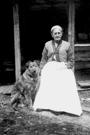Late 1800's Old Woman Sitting Outside Wood Cabin Wearing Dress With Beautiful Dog Sitting Beside Her