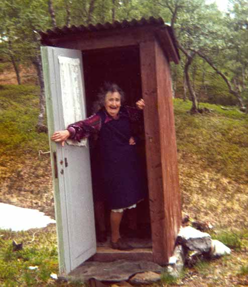 Older Woman Standing Inside Outhouse Toilet Building With Arm On Open Door European Countryside With Trees